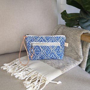 May include: A blue and white patterned clutch with a zipper closure and a leather strap. The clutch is resting on a beige and white fringed blanket. The background includes a beige chair and a green plant.