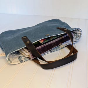May include: A blue tote bag with brown leather handles and accents. The bag is open, revealing a floral patterned lining and several books and papers inside. The bag is made of a sturdy material and has a button closure.