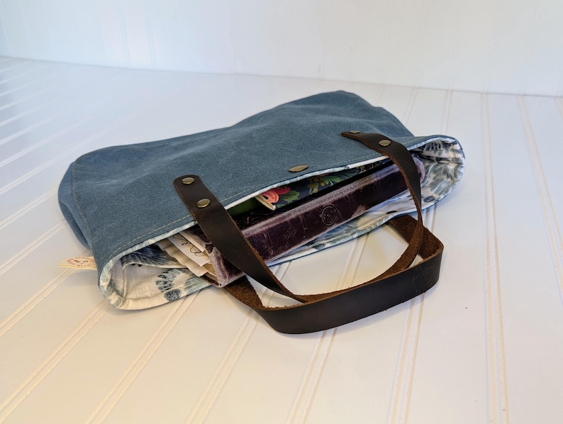 May include: A blue tote bag with brown leather handles and accents. The bag is open, revealing a floral patterned lining and several books and papers inside. The bag is made of a sturdy material and has a button closure.