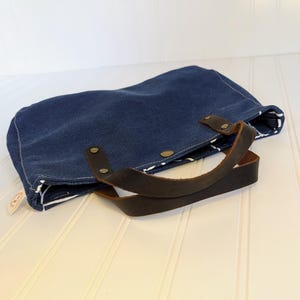 May include: A navy blue tote bag with brown leather handles and accents. The bag is made of a sturdy fabric and features a white and black patterned lining. The handles are attached with brass rivets.