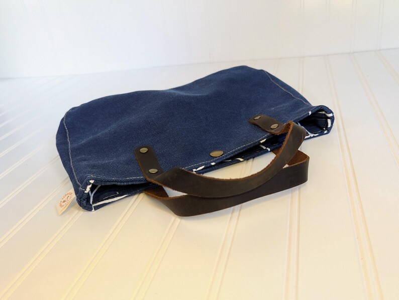 May include: A navy blue tote bag with brown leather handles and accents. The bag is made of a sturdy fabric and features a white and black patterned lining. The handles are attached with brass rivets.