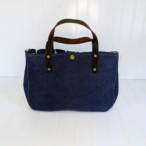 May include: A navy blue denim tote bag with brown leather handles and brass rivets. The bag has a rectangular shape with a rounded bottom and a button closure. The interior lining is patterned.