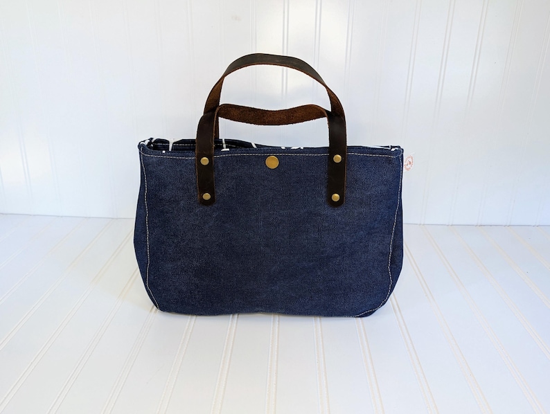 May include: A navy blue denim tote bag with brown leather handles and brass rivets. The bag has a rectangular shape with a rounded bottom and a button closure. The interior lining is patterned.