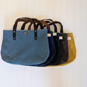May include: A row of five canvas tote bags in various colors: blue, navy, charcoal, and mustard yellow. Each bag has brown leather handles and a snap closure. The bags are displayed against a white, vertical-slat background.
