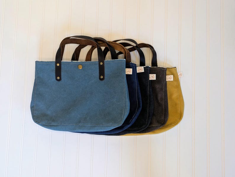 May include: A row of five canvas tote bags in various colors: blue, navy, charcoal, and mustard yellow. Each bag has brown leather handles and a snap closure. The bags are displayed against a white, vertical-slat background.