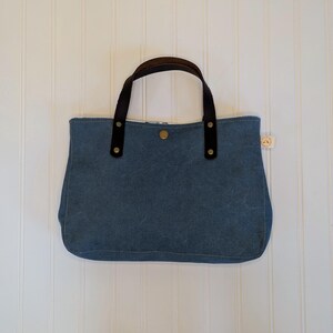 May include: A blue tote bag with brown leather handles and accents. The bag has a button closure and a small tag on the side. The bag is made of a sturdy material and is designed for carrying items.