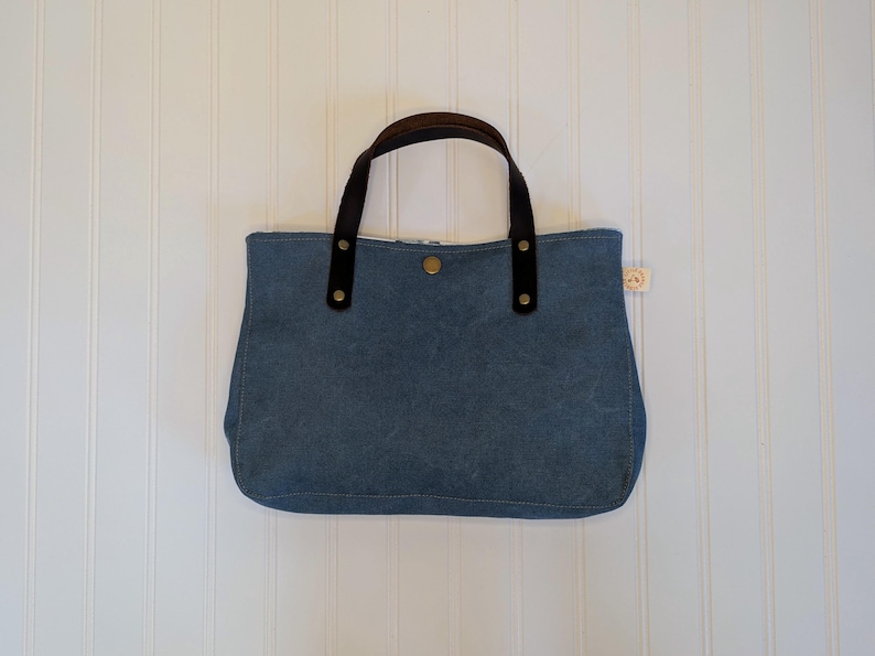 May include: A blue tote bag with brown leather handles and accents. The bag has a button closure and a small tag on the side. The bag is made of a sturdy material and is designed for carrying items.