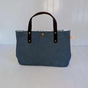 May include: A blue tote bag with brown leather handles and accents. The bag has a rectangular shape with a button closure and a small tag. The handles are attached with rivets. The interior lining is visible.