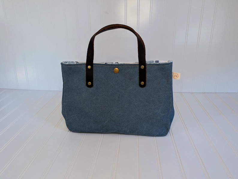May include: A blue tote bag with brown leather handles and accents. The bag has a rectangular shape with a button closure and a small tag. The handles are attached with rivets. The interior lining is visible.
