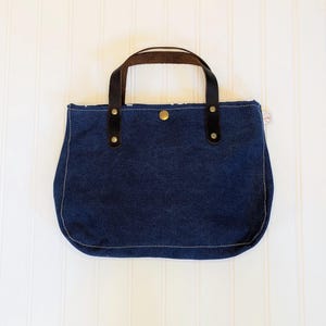 May include: A navy blue tote bag with brown leather handles and black accents. The bag has a rounded bottom and is secured with a snap closure. The stitching is a light cream color.
