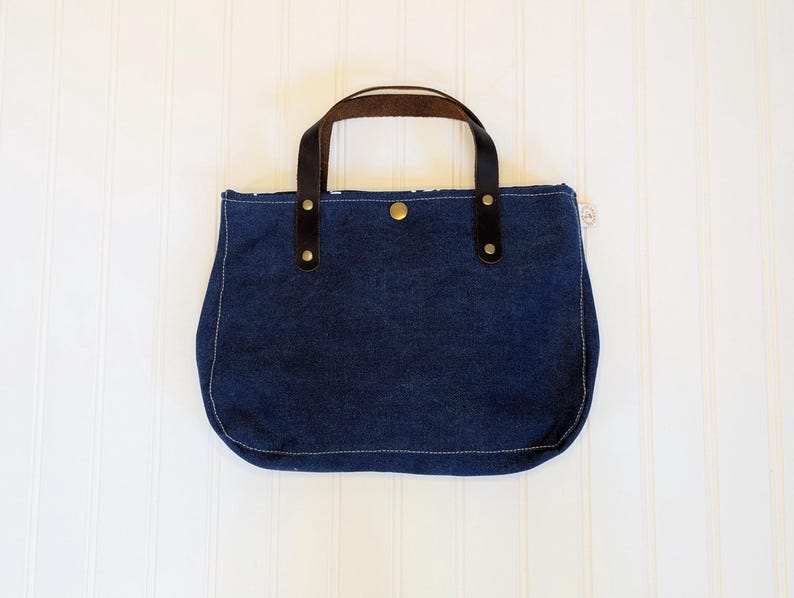 May include: A navy blue tote bag with brown leather handles and black accents. The bag has a rounded bottom and is secured with a snap closure. The stitching is a light cream color.