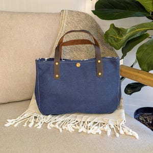 May include: A blue denim tote bag with brown leather handles and brass rivets. The bag is rectangular with a rounded bottom and a patterned interior. It sits on a beige blanket with white fringe.