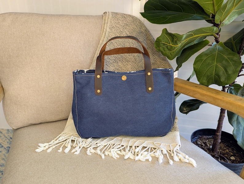 May include: A blue denim tote bag with brown leather handles and brass rivets. The bag is rectangular with a rounded bottom and a patterned interior. It sits on a beige blanket with white fringe.