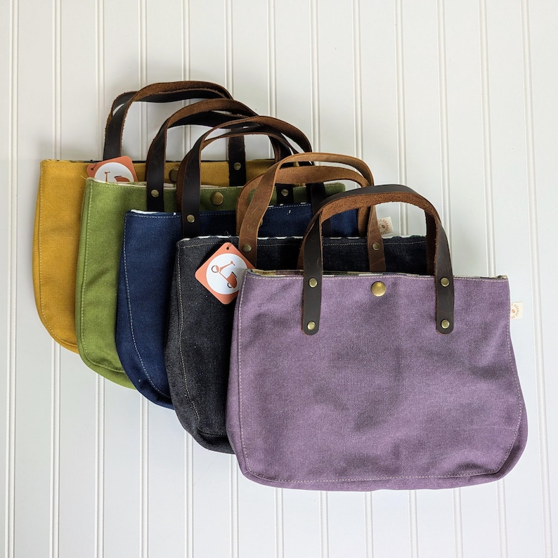 May include: A row of five canvas tote bags in various colors: mustard yellow, olive green, navy blue, charcoal gray, and lavender. Each bag has brown leather handles and a button closure. A tag is attached to one bag.