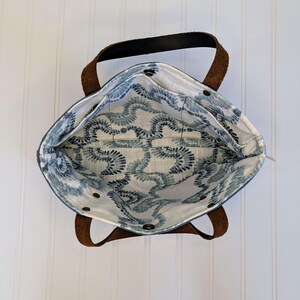 May include: An overhead view of a tote bag with brown leather handles and a black top handle. The interior features a blue and white patterned fabric. The bag is open, revealing the inside pockets and the patterned lining. The bag is sitting on a white surface.