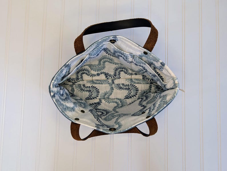 May include: An overhead view of a tote bag with brown leather handles and a black top handle. The interior features a blue and white patterned fabric. The bag is open, revealing the inside pockets and the patterned lining. The bag is sitting on a white surface.