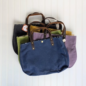 May include: A collection of canvas tote bags in various colors, including navy blue, olive green, and purple. Each bag features brown leather handles and a button closure. The bags are displayed against a white background.