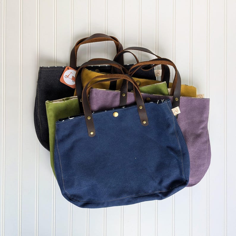 May include: A collection of canvas tote bags in various colors, including navy blue, olive green, and purple. Each bag features brown leather handles and a button closure. The bags are displayed against a white background.