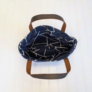 May include: A navy blue tote bag with a white geometric pattern on the interior lining. The bag has brown leather handles and a simple, open design. The bag is sitting on a white surface.