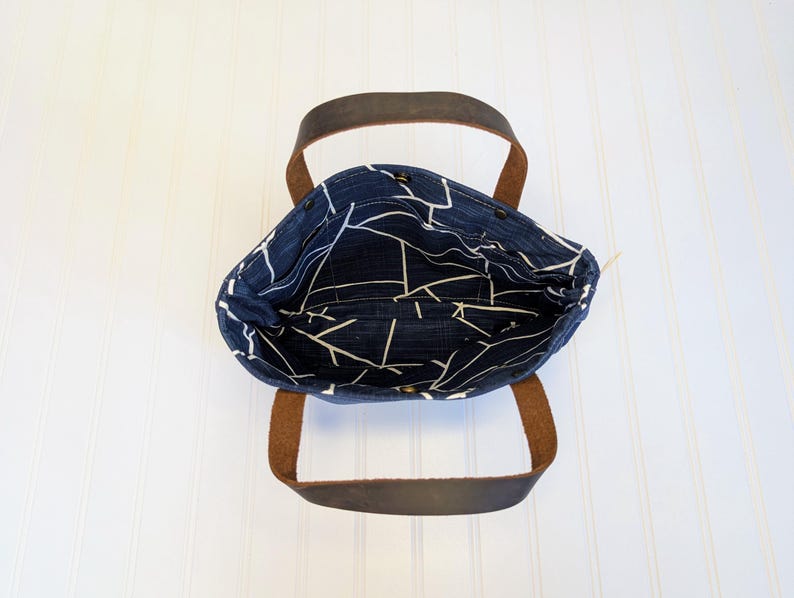 May include: A navy blue tote bag with a white geometric pattern on the interior lining. The bag has brown leather handles and a simple, open design. The bag is sitting on a white surface.