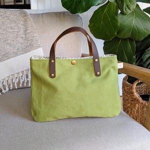 May include: A green canvas tote bag with brown leather handles and brass rivets. The bag has a button closure and a patterned interior. The tote is sitting on a gray chair next to a woven basket and a fringed blanket.