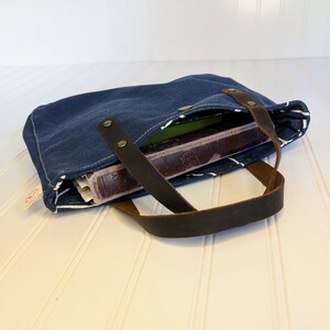 May include: A blue denim tote bag with brown leather handles. The bag is open, revealing a book and other items inside. The bag has white stitching and a striped lining.