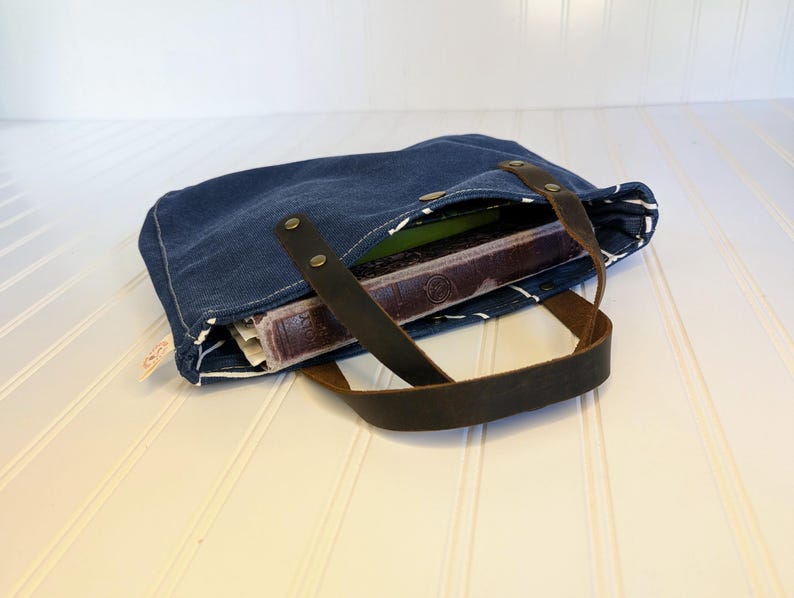 May include: A blue denim tote bag with brown leather handles. The bag is open, revealing a book and other items inside. The bag has white stitching and a striped lining.