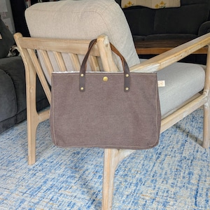 May include: A brown canvas tote bag with leather handles and brass rivets. The bag is rectangular with a button closure and is sitting on a wooden chair. The background includes a blue rug and a neutral-toned chair.