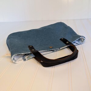 May include: A blue tote bag with a dark brown leather handle. The bag has a floral patterned lining and is secured with a snap closure. The bag is made of a sturdy material.