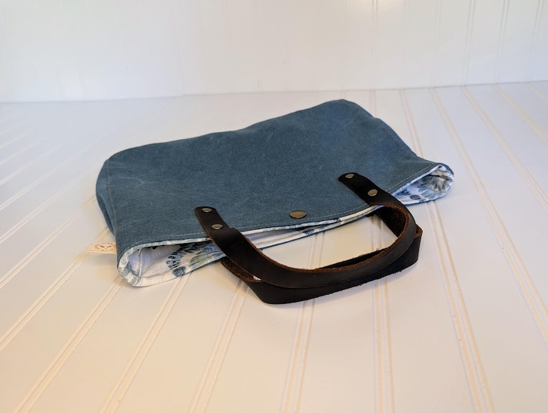 May include: A blue tote bag with a dark brown leather handle. The bag has a floral patterned lining and is secured with a snap closure. The bag is made of a sturdy material.