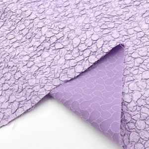May include: A close-up of a lilac textured fabric with a crinkled, pebble-like pattern. The fabric is folded over, revealing a lighter lilac lining with a subtle pattern.