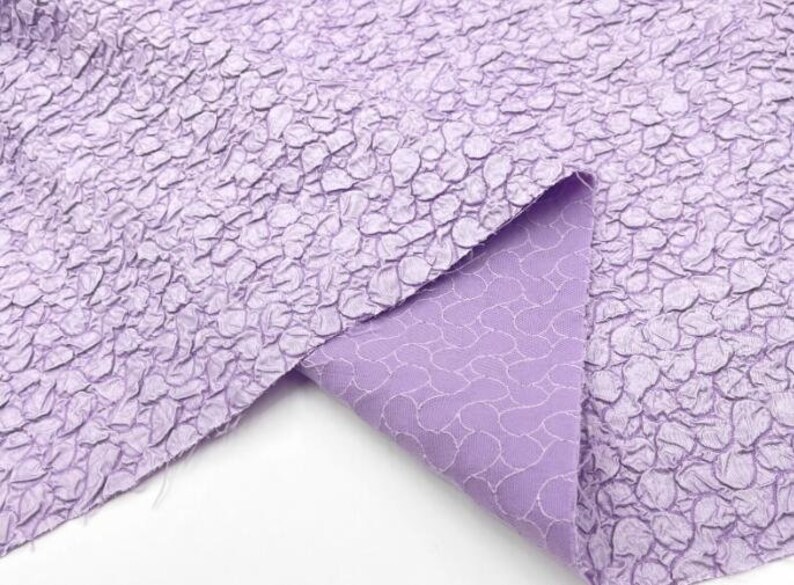 May include: A close-up of a lilac textured fabric with a crinkled, pebble-like pattern. The fabric is folded over, revealing a lighter lilac lining with a subtle pattern.