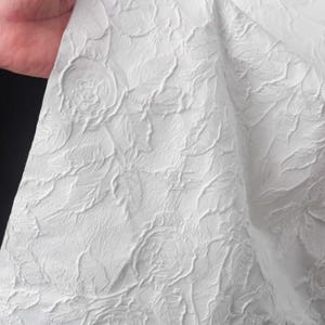 Pode incluir: Close-up de tecido branco com um padrão floral em relevo. O tecido é segurado por uma mão, revelando a textura e o design. O padrão apresenta formas detalhadas de rosas e linhas fluidas, criando uma superfície texturizada.
