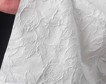 May include: A close-up of white fabric with a raised floral pattern. The fabric is held by a hand, revealing the texture and design. The pattern features detailed rose-like shapes and flowing lines, creating a textured surface.