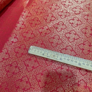 Könnte beinhalten: Roter Stoff mit einem goldenen, metallischen geometrischen Muster. Der Stoff wird mit einem Lineal gemessen, auf dem "Made in China" steht.