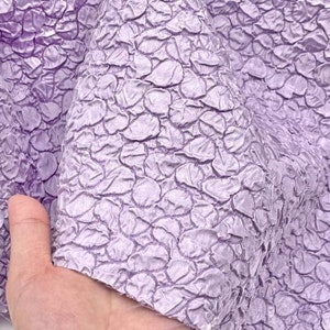 May include: A close-up of a lilac textured fabric with a crinkled, bubble-like pattern. The fabric is soft and has a subtle sheen.