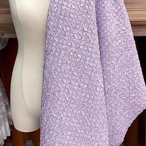 May include: A lilac textured fabric with a raised pattern of small circles. The fabric is draped over a white dress form.