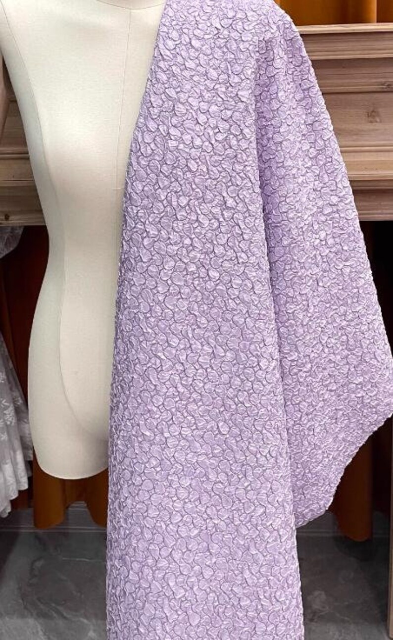 May include: A lilac textured fabric with a raised pattern of small circles. The fabric is draped over a white dress form.