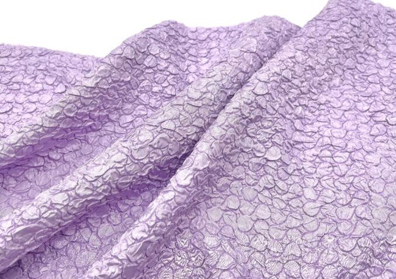 May include: A close-up of a textured, lilac fabric with a raised pattern. The fabric is soft and has a subtle sheen.