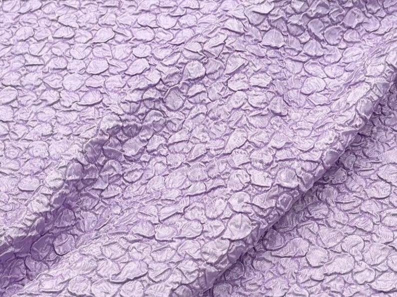 May include: Close-up of a textured fabric with a light purple color. The fabric has a raised, pebble-like pattern.