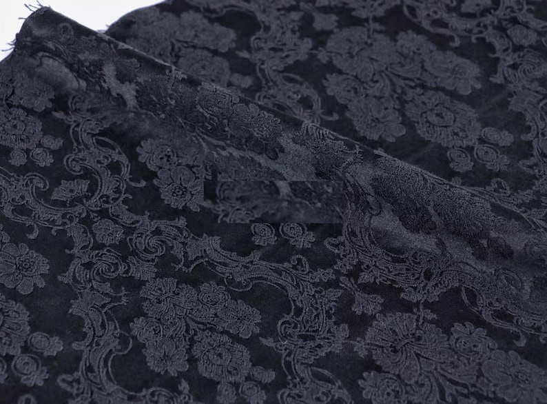 Pode incluir: Tecido azul marinho escuro com um padr&atilde;o floral e de rolagem detalhado. O tecido est&aacute; dobrado, mostrando a textura e profundidade do design. O padr&atilde;o &eacute; em um tom mais claro de azul marinho, criando um contraste sutil.