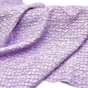 May include: A close-up of a textured, lilac fabric with a pebble-like pattern. The fabric is folded and draped, showcasing its soft and crinkled texture.