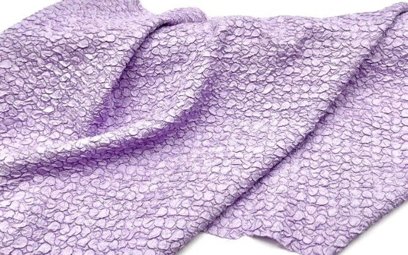 May include: A close-up of a textured, lilac fabric with a pebble-like pattern. The fabric is folded and draped, showcasing its soft and crinkled texture.