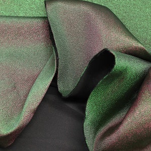 May include: Close-up of a green and iridescent fabric with a textured surface. The fabric is folded and draped, showing the shimmery effect of the material.