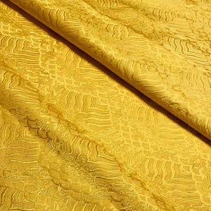 Pode incluir: Close-up de um tecido amarelo dobrado com uma superfície texturizada. O material apresenta um padrão intrincado de linhas onduladas e formas de escamas, criando uma superfície rica e tátil. As dobras do tecido adicionam profundidade e dimensão.