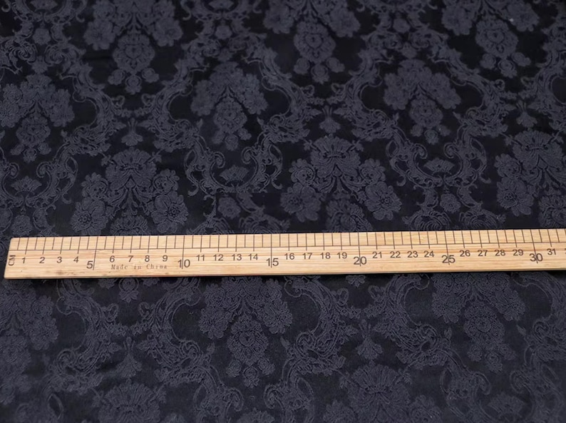 Pode incluir: Tecido escuro com um padr&atilde;o floral repetido. Uma r&eacute;gua de madeira, marcada em cent&iacute;metros, est&aacute; sobre o tecido, medindo at&eacute; 79 cm. O tecido tem um brilho sutil e um design detalhado e texturizado.