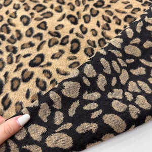 May include: A close-up of a fabric with a leopard print pattern. The fabric is brown with black spots. The fabric is folded in half, showing both sides of the fabric.