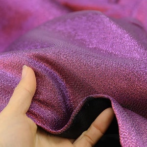 May include: Close-up of a shimmering purple and pink fabric with a subtle iridescent sheen. The fabric has a textured, woven appearance.