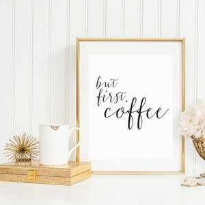 May include: A gold framed print with the text "but first, coffee" in black cursive script. The print is on a white background and is sitting on a white surface. There is a white mug and a gold decorative object in the foreground.