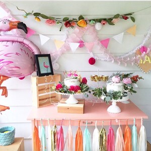 May include: A pink flamingo balloon, a wooden crate, and two white cakes decorated with flowers and greenery on a pink table with a colorful tassel garland. The table is set up for a party with a pink and green theme.